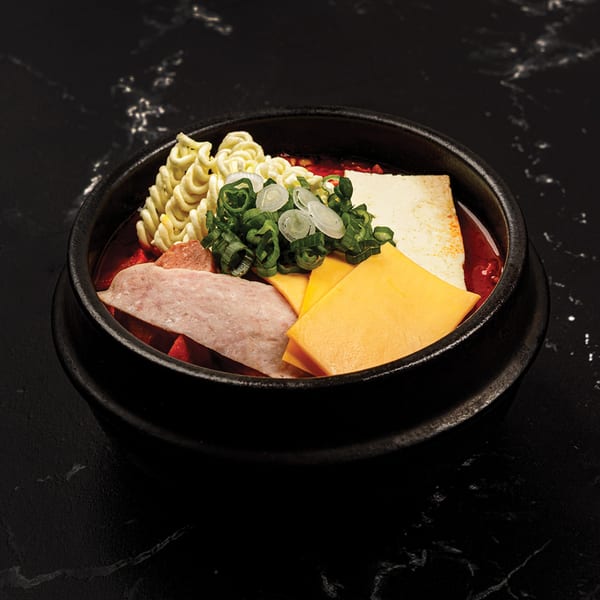 Antoya Korean BBQ's Budae Jjigae with ramen, tofu, ham, cheese, and scallions in a black bowl.