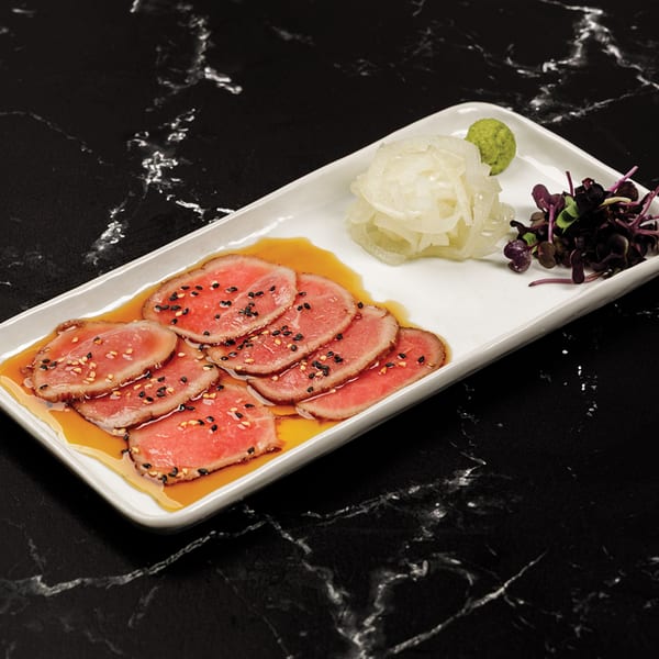 Thinly sliced beef in sauce with sesame seeds, pickled onions, wasabi, and microgreens on a white plate.
